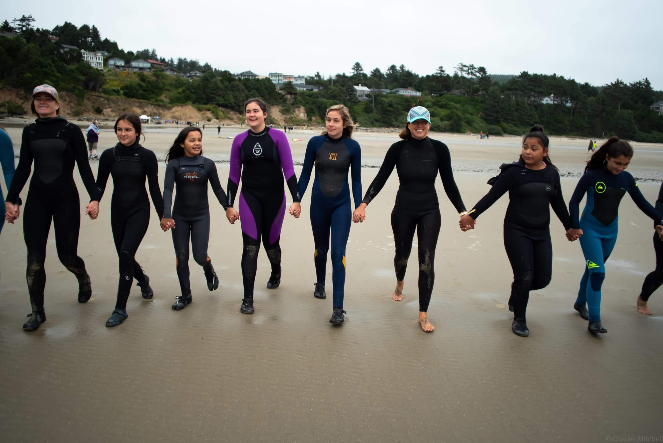 Women and girls in wetsuits holding hands and walking on the beach together.