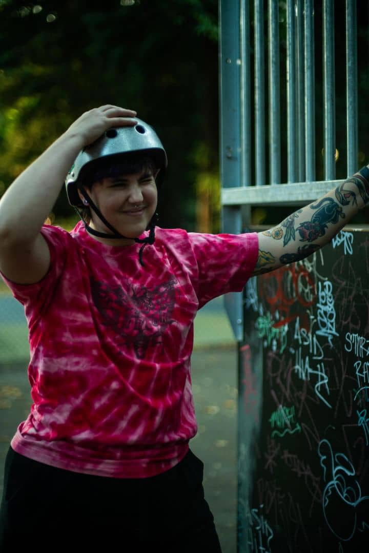 Alex Gray chilling at the skate park wearing a tie-dye GNDR SHREDR shirt. They have their hand on top of their helmet with a scrunched up smile.